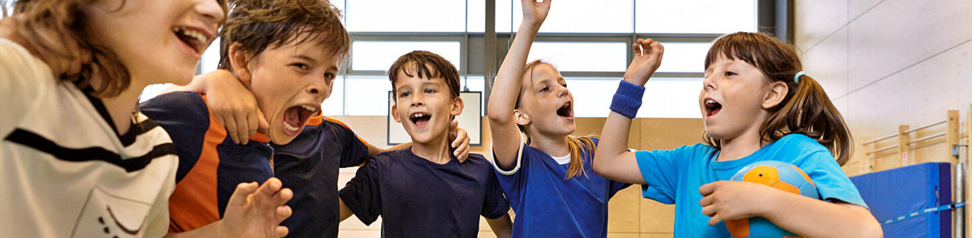 Kinder in verschiedenen Sport-Outfits fallen sich lachend in die Arme.