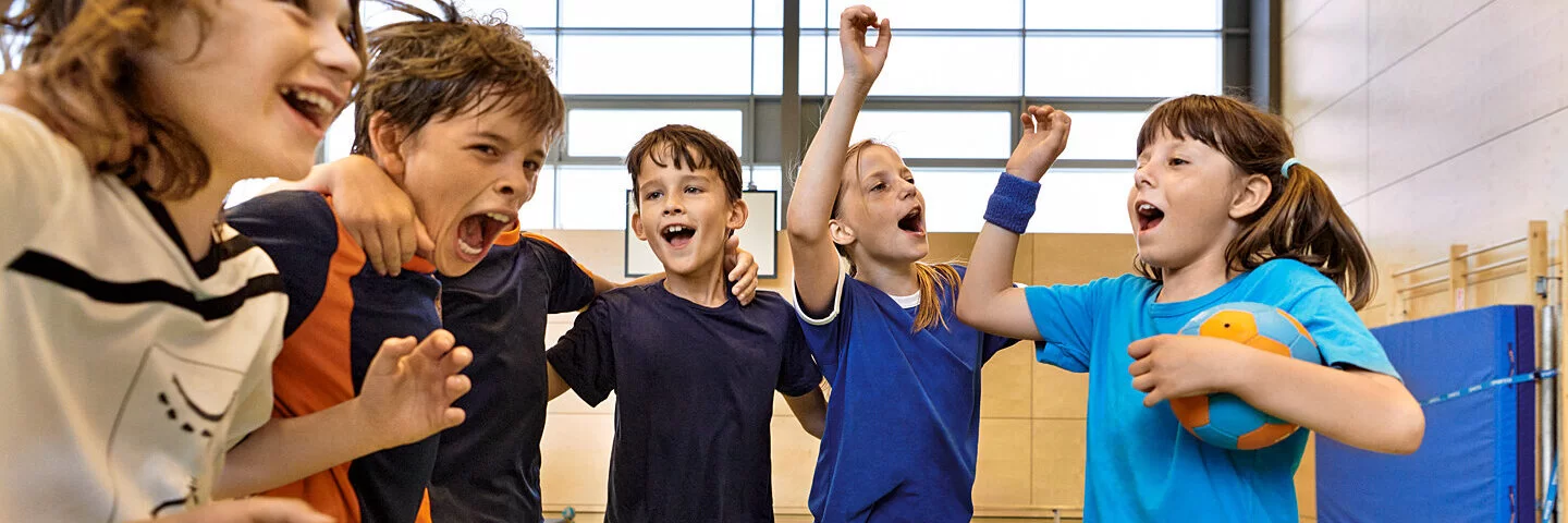 Kinder in verschiedenen Sport-Outfits fallen sich lachend in die Arme.