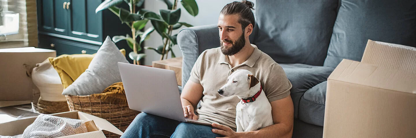 Ein junger Mann sitzt mit seinem Hund auf dem Boden und schaut auf sein Laptop.