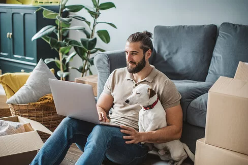 Ein junger Mann sitzt mit seinem Hund auf dem Boden und schaut auf sein Laptop.