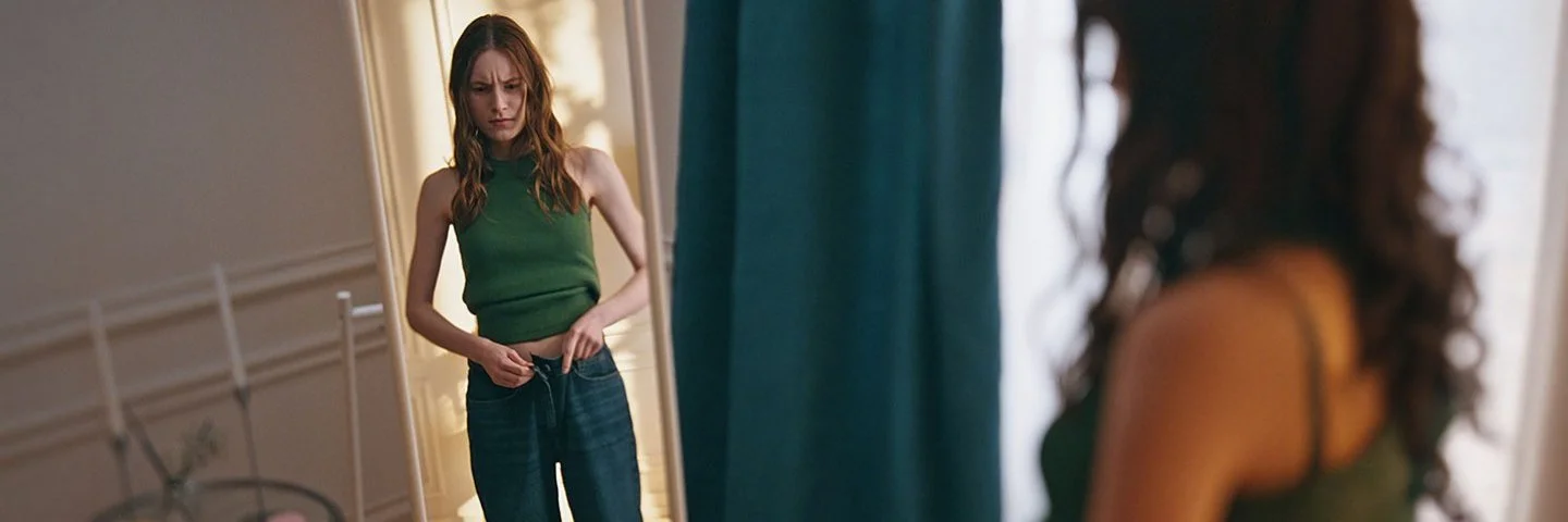 Eine junge magere Frau steht vor einem Spiegel und betrachtet sich kritisch.