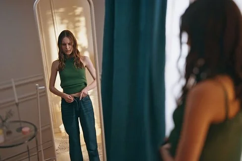 Eine junge magere Frau steht vor einem Spiegel und betrachtet sich kritisch.