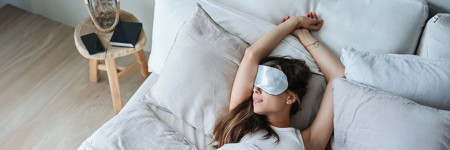 Eine junge Frau iegt mit einer Schlafmaske im Bett und streckt die Arme über den Kopf.