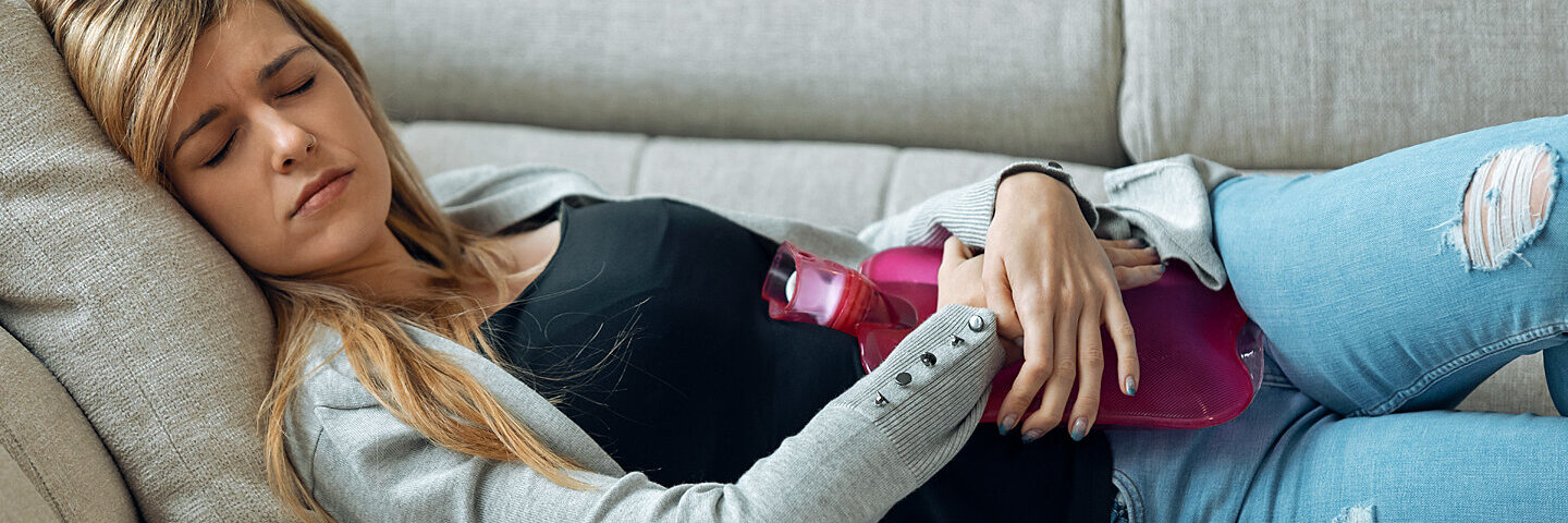 Junge Frau mit einer Blasenentzündung liegt auf dem Sofa und wärmt sich mit einer Wärmflasche.