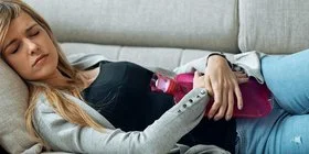 Junge Frau mit einer Blasenentzündung liegt auf dem Sofa und wärmt sich mit einer Wärmflasche.
