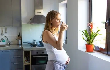 Eine schwangere Frau steht vor einem Fenster und trinkt ein Glas Wasser.