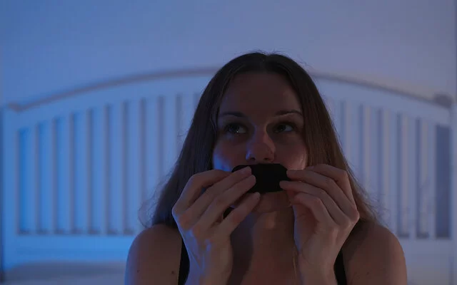 Eine junge Frau sitzt auf ihrem Bett und klebt sich ein Pflaster auf den Mund.