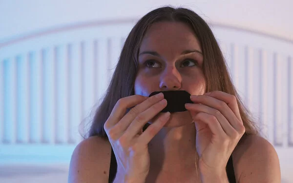 Eine junge Frau sitzt auf ihrem Bett und klebt sich ein Pflaster auf den Mund.