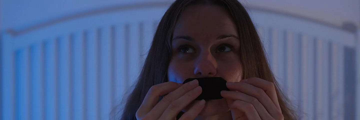 Eine junge Frau sitzt auf ihrem Bett und klebt sich ein Pflaster auf den Mund.