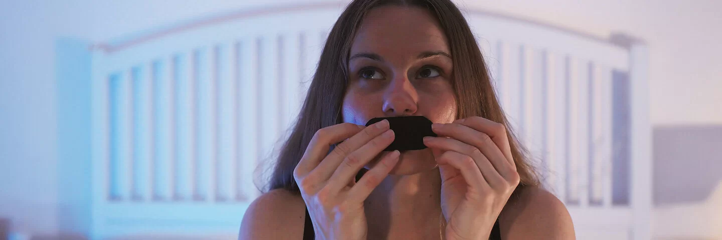 Eine junge Frau sitzt auf ihrem Bett und klebt sich ein Pflaster auf den Mund.