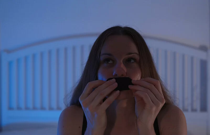 Eine junge Frau sitzt auf ihrem Bett und klebt sich ein Pflaster auf den Mund.