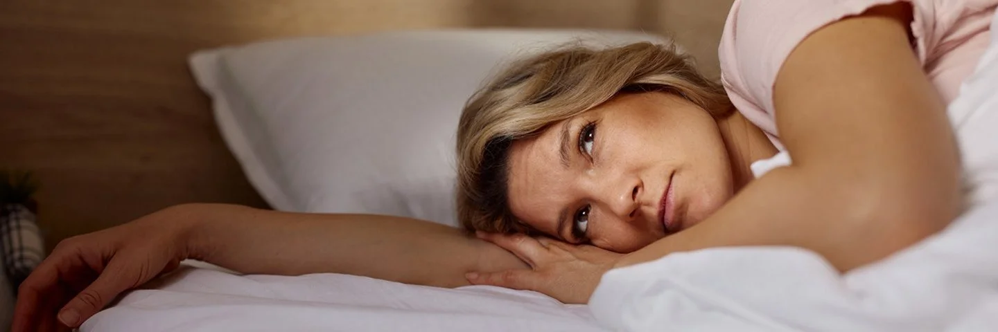 Eine Frau liegt mit geöffneten Augen auf der Seite im Bett unter einer Decke. Ihr Kopf ruht auf dem ausgestreckten rechten Arm und ihr Blick geht nach oben.