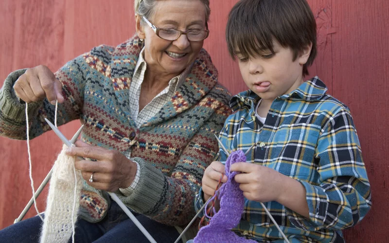 Eine ältere Frau mit Stricksachen sitzt neben einem Jungen und zeigt ihm das Stricken.