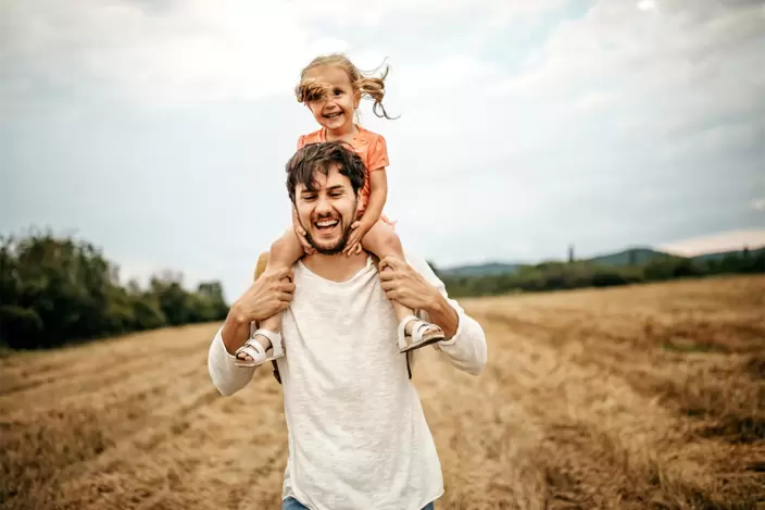 Ein junger Vater trägt seine Tochter auf den Schultern über ein Feld.