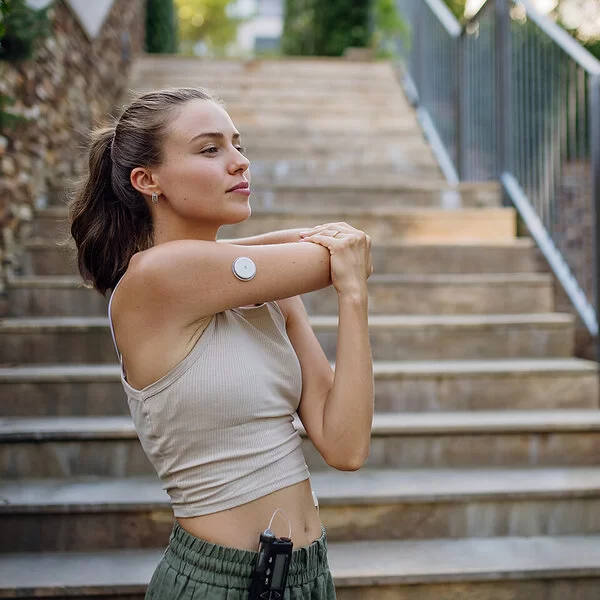 Eine junge Frau in sportlicher Kleidung. An ihrem rechten Oberarm ist ein Sensor zum Blutzuckermessen sichtbar.