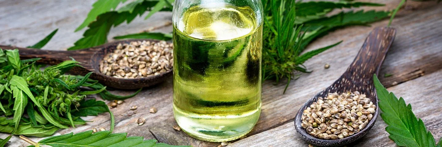 Hanfsamen in Holzlöffeln und Öl in einer offenen Glasflasche auf einem rustikalem Holztisch dekoriert mit Hanfblättern.