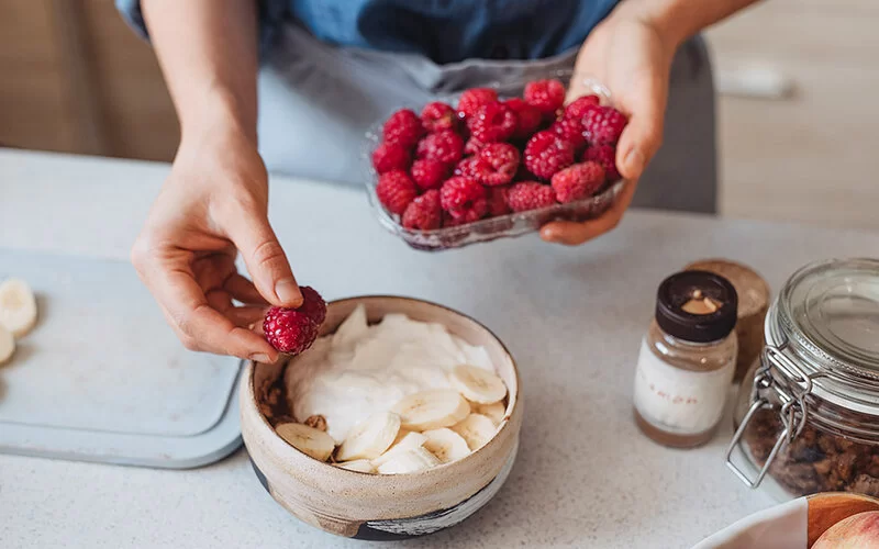 Eine Frau gibt frische Himbeeren in eine Schüssel mit Bananen und Joghurt.
