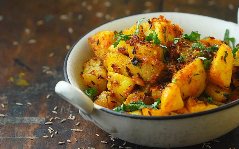 Auf einem Holztisch steht eine Schale mit dem traditionellen indischen Kartoffelgericht Jeera Aloo. Die angebratenen Kartoffeln sind mit Kräutern und Kreuzkümmelsamen bestreut. Weitere Samen liegen verstreut um die Schüssel herum auf dem Tisch.