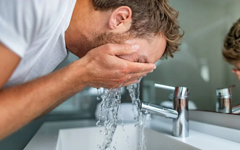 Ein Mann beugt sich über einen Waschtisch und hält die Hände ins Gesicht, Wasser spritzt durch seine Finger.