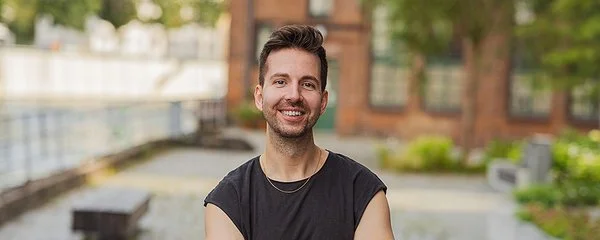 Tobias Herrmann-Schwarz trägt ein kurzärmeliges T-Shirt und steht mit verschränkten Armen im Freien vor einem Backsteingebäude.