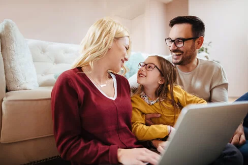 Eine junge Familie sitzt gemeinsam vor ihrem Sofa am Laptop und lacht.