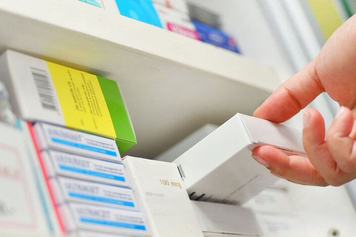 Ein Regal in einer Apotheke, in dem mehrere Medikamentenpackungen liegen. Eine Person greift eines der Medikamente und zieht es aus dem Regal.