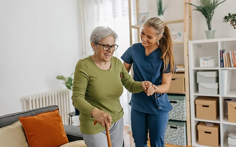 Eine Pflegefachperson hilft einer älteren Dame mit Gehilfe vom Sofa auf.
