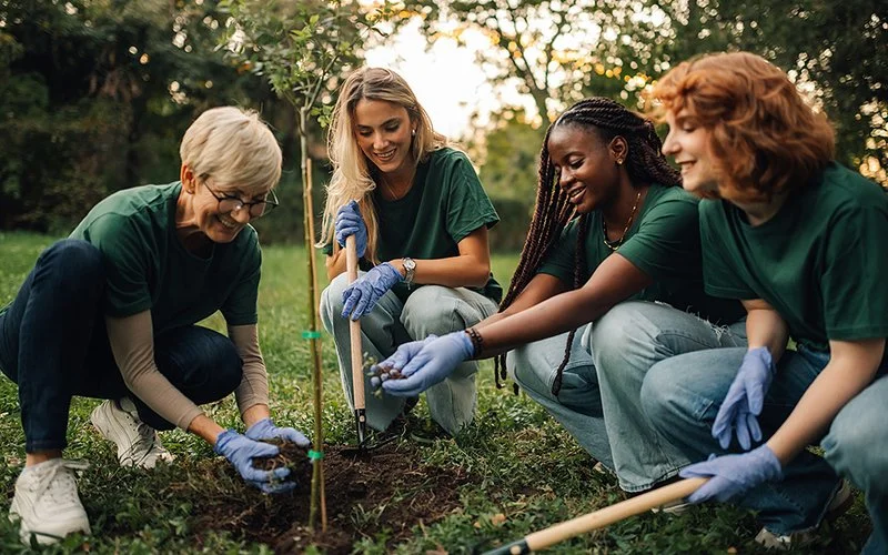 Vier Frauen verschiedenen Alters pflanzen in einem Park gemeinsam einen Baumsetzling. Sie tragen grüne T-Shirts und blaue Arbeitshandschuhe. Der Baum befindet sich bereits im Pflanzloch, um das die Frauen knien. Eine von ihnen hält eine Schaufel, eine andere eine Hacke in der Hand. Die beiden anderen füllen mit ihren Händen Erde in das Loch.