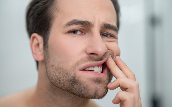 Ein junger Mann schaut kritisch in den Spiegel, schiebt mit der linken Hand die Lippe etwas hoch und schaut, ob er eine Zahnfleischentzündung hat.