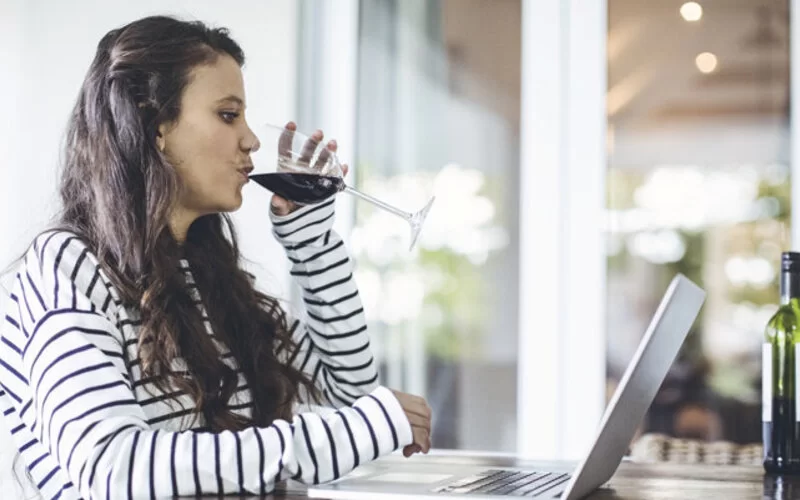 Eine Frau sitzt am Tisch vor dem Laptop und trinkt Rotwein.