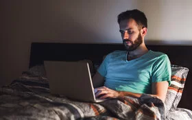 Ein Mann sitzt mit angeschaltetem Laptop im Bett, das davon ausgehende blaue Licht könnte seinen Schlaf negativ beeinträchtigen.