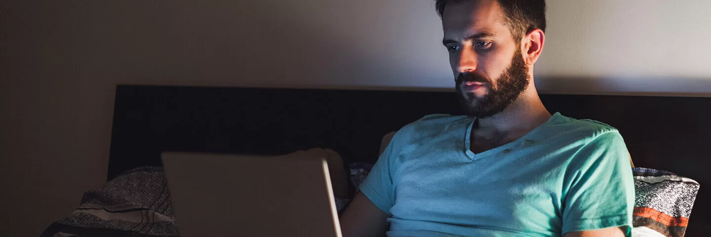 Ein Mann sitzt mit angeschaltetem Laptop im Bett, das davon ausgehende blaue Licht könnte seinen Schlaf negativ beeinträchtigen.