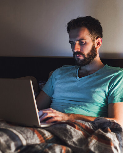 Ein Mann sitzt mit angeschaltetem Laptop im Bett, das davon ausgehende blaue Licht könnte seinen Schlaf negativ beeinträchtigen.
