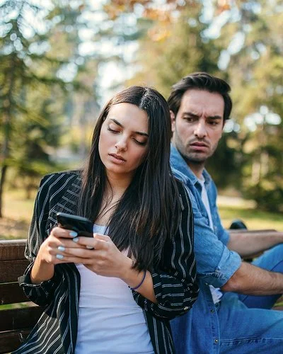 Ein Mann und eine Frau sitzen auf einer Bank im Park. Sie dreht ihm den Rücken zu und schaut auf ihr Handy. Er blickt wütend zu ihr rüber.