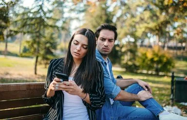 Ein Mann und eine Frau sitzen auf einer Bank im Park. Sie dreht ihm den Rücken zu und schaut auf ihr Handy. Er blickt wütend zu ihr rüber.