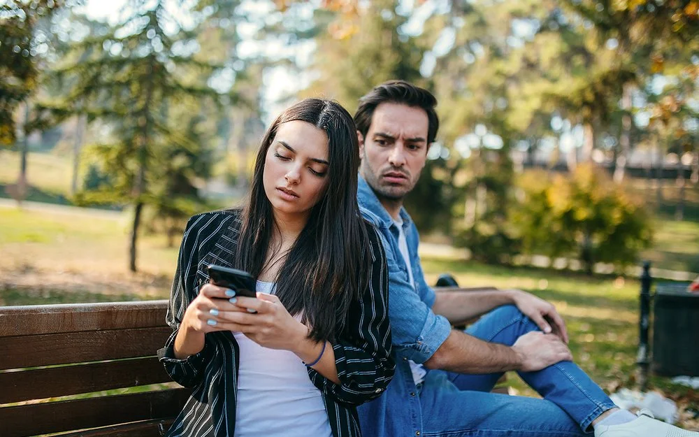 Ein Mann und eine Frau sitzen auf einer Bank im Park. Sie dreht ihm den Rücken zu und schaut auf ihr Handy. Er blickt wütend zu ihr rüber.