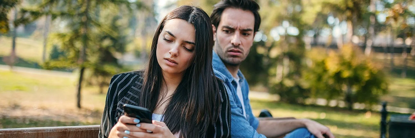 Ein Mann und eine Frau sitzen auf einer Bank im Park. Sie dreht ihm den Rücken zu und schaut auf ihr Handy. Er blickt wütend zu ihr rüber.