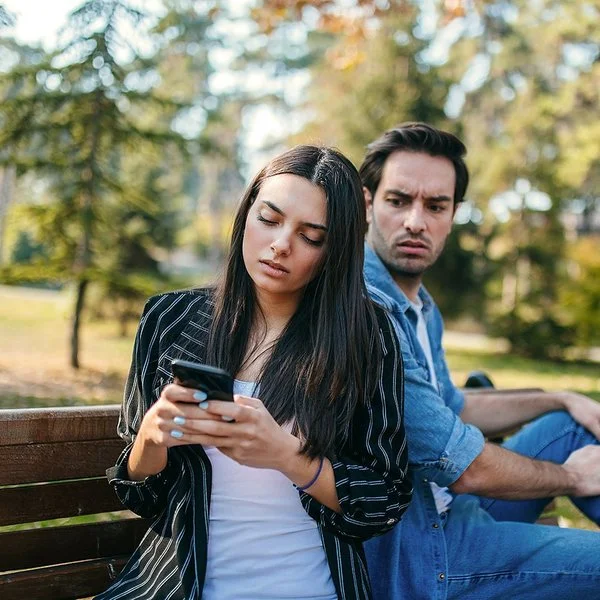Ein Mann und eine Frau sitzen auf einer Bank im Park. Sie dreht ihm den Rücken zu und schaut auf ihr Handy. Er blickt wütend zu ihr rüber.