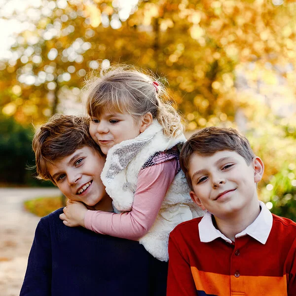 Drei Geschwister, zwei Jungen und ein Mädchen, halten sich in einem Park auf. Im Hintergrund befinden sich herbstlich gefärbte Laubbäume. Das Mädchen ist das jüngste der drei und umarmt den ältesten Bruder.