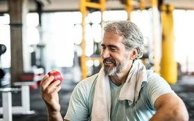 Ein Mann Mitte 50 in Sportkleidung und einem Handtuch um den Hals isst in einem Fitnessstudio einen Apfel.