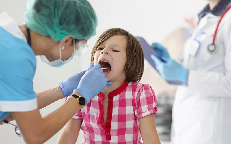 Eine Ärztin mit medizinischer Maske untersucht wegen Verdacht auf Mandelentzündung den Rachen eines Mädchens mit einem Spatel.