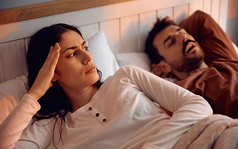 Ein Paar liegt nebeneinander im Bett. Der Mann schläft mit offenem Mund. Die Frau ist wach, stützt den Kopf in die Hand und schaut ernst zu dem Mann herüber..