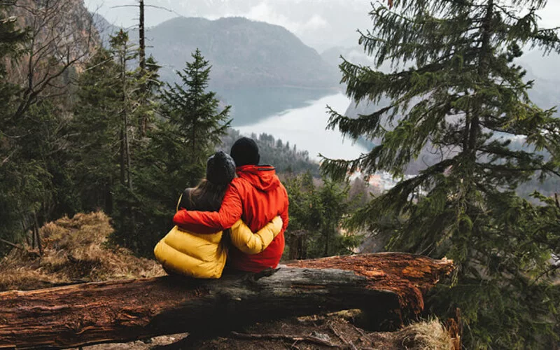 Ein Mann und eine Frau sitzen auf einem Baumstamm in einer Berglandschaft und umarmen sich. Sie schauen auf ein Tal und einen See.