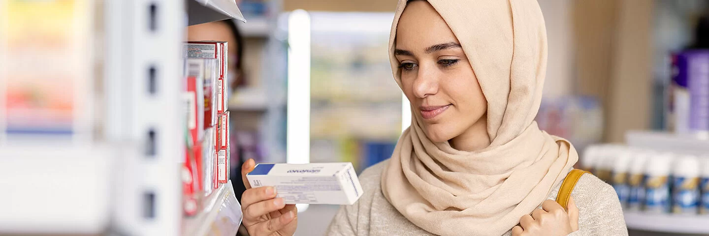 Eine junge Frau mit Hijab steht in einer Apotheke und liest die Aufschrift einer Medikamentenverpackung.
