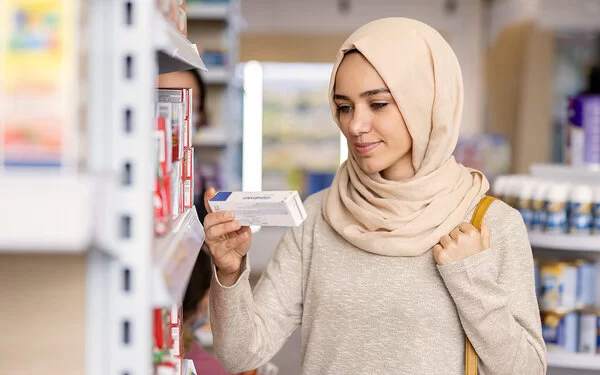 Eine junge Frau mit Hijab steht in einer Apotheke und liest die Aufschrift einer Medikamentenverpackung.
