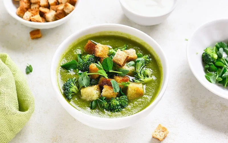 Nahaufnahme einer Brokkolisuppe mit Croutons in einer Schüssel, die auf hellem Steinhintergrund platziert ist.