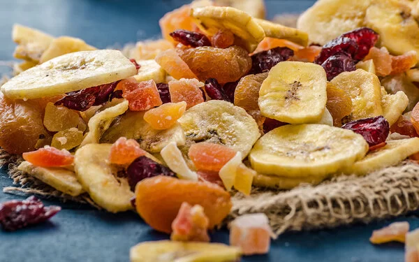 Eine Mischung von Trockenfrüchten mit Bananenchips, Aprikosen, Preiselbeeren und Papaya auf einer dunkelgrauen Tischplatte.