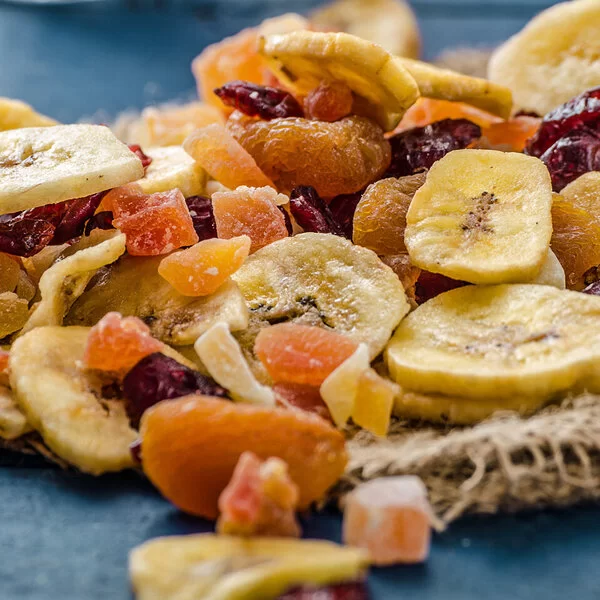 Eine Mischung von Trockenfrüchten mit Bananenchips, Aprikosen, Preiselbeeren und Papaya auf einer dunkelgrauen Tischplatte.