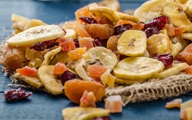 Eine Mischung von Trockenfrüchten mit Bananenchips, Aprikosen, Preiselbeeren und Papaya auf einer dunkelgrauen Tischplatte.