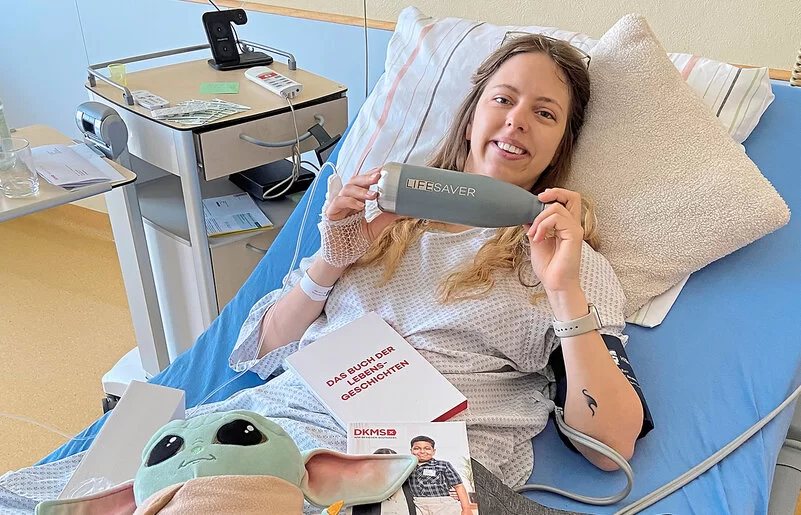 Eine Frau liegt in einem Krankenhausbett. Sie lächelt und hält mit beiden Händen eine Trinkflasche hoch, auf der Lifesaver steht.